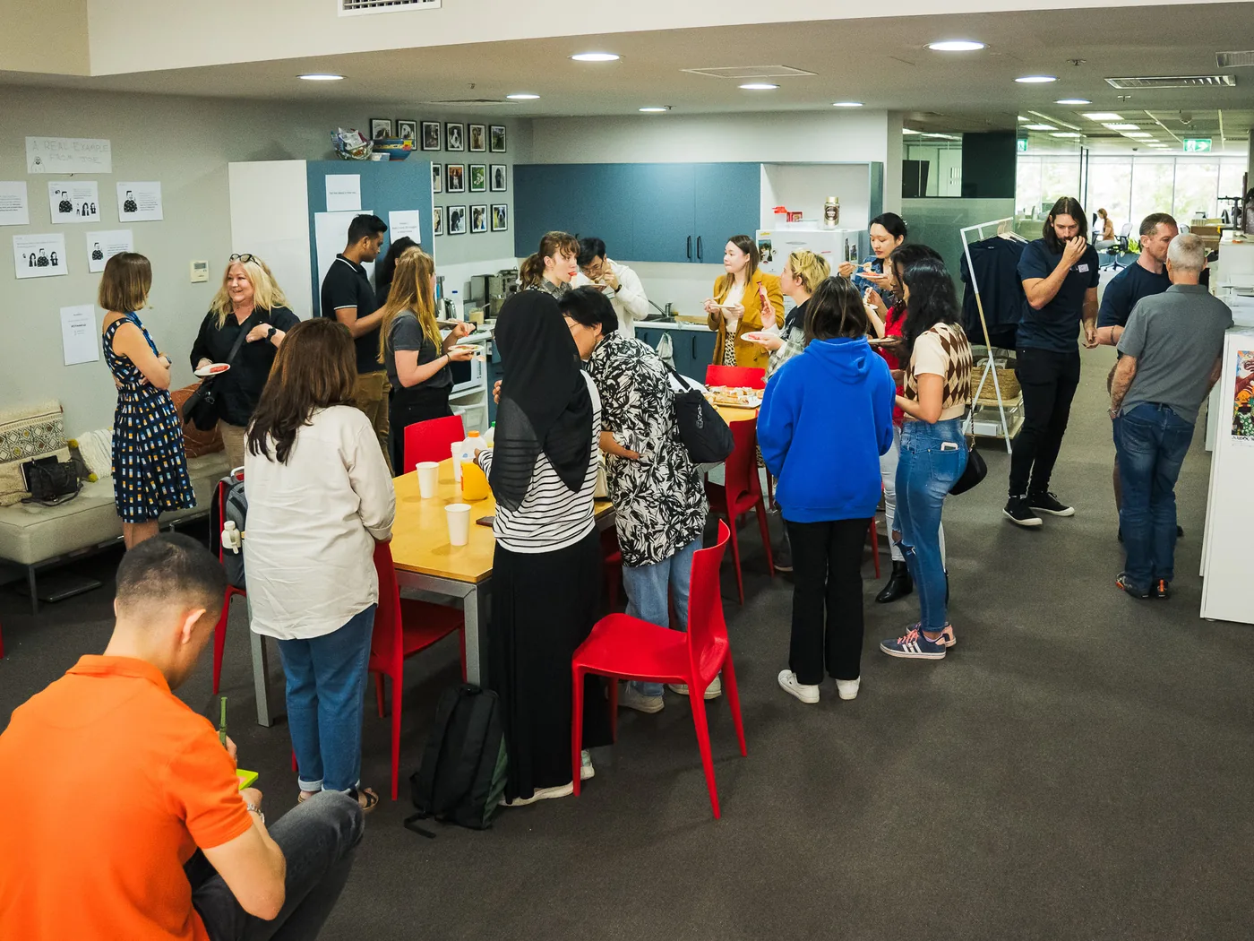 People standing and talking around tables in a shared office kitchen, printed sheets pinned to the wall behind them.